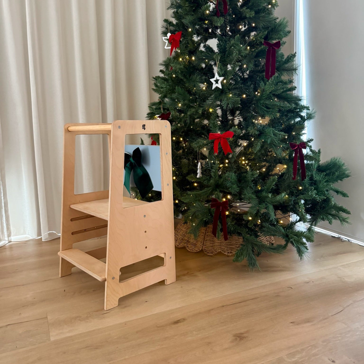 Wooden kitchen helper in front of a decorated Christmas tree with red ribbons on a wooden floor.