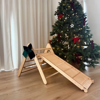 Wooden pikler ramp with a Christmas tree decorated with red bows in a room.
