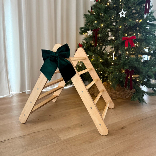 Wooden pilker climbing frame with a green bow against a Christmas tree decorated with red and gold ornaments.